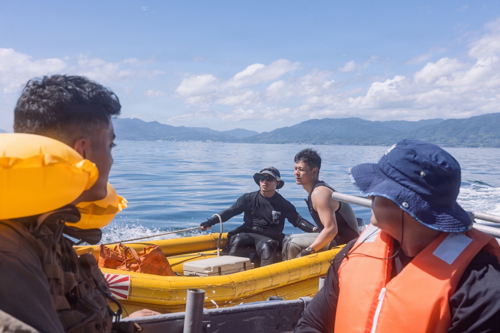 The image shows a group of people on the water, with two individuals in a yellow inflatable rescue/utility boat being towed or connected by rope to a larger vessel. The scene appears to take place on a calm bay or sea with mountainous, forested coastline visible in the background. Some individuals are wearing life jackets, suggesting a maritime safety or rescue-related activity.