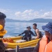 The image shows a group of people on the water, with two individuals in a yellow inflatable rescue/utility boat being towed or connected by rope to a larger vessel. The scene appears to take place on a calm bay or sea with mountainous, forested coastline visible in the background. Some individuals are wearing life jackets, suggesting a maritime safety or rescue-related activity.