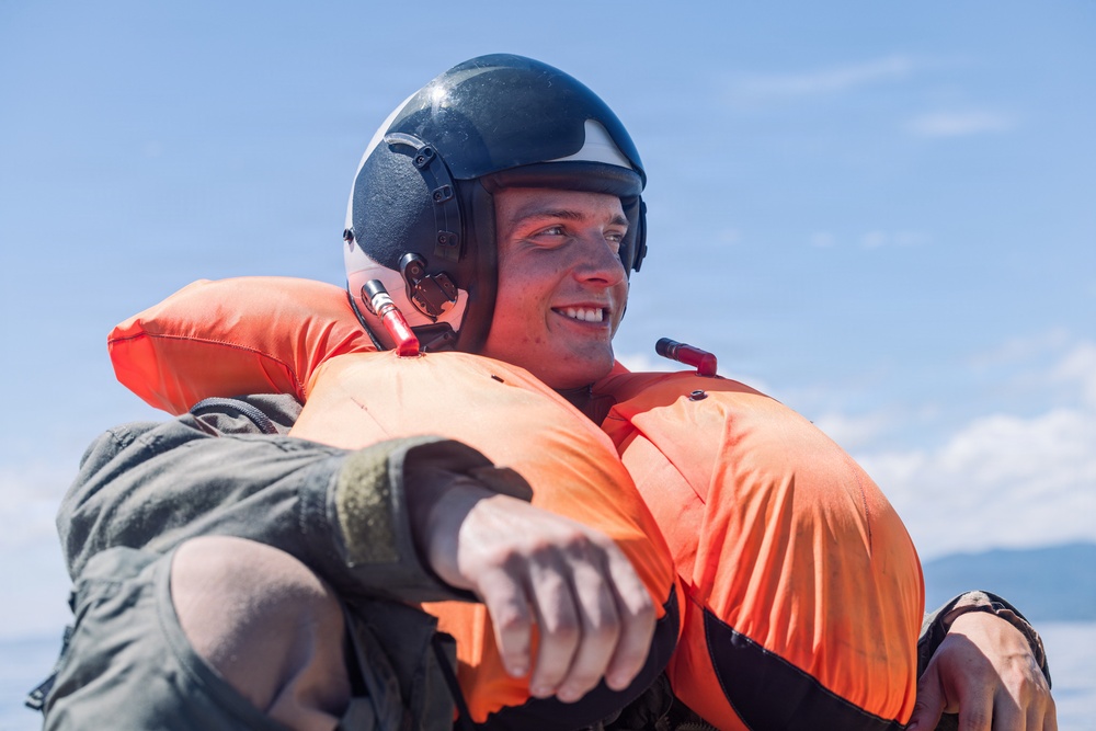 A young male wearing a dark flight helmet with communications equipment and an inflated orange life preserver vest is smiling while carrying or supporting another person. The setting appears to be outdoors under a clear blue sky, suggesting a water rescue or military aviation survival training scenario. The subject appears confident and in good spirits despite what appears to be a demanding physical situation.