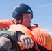 A young male wearing a dark flight helmet with communications equipment and an inflated orange life preserver vest is smiling while carrying or supporting another person. The setting appears to be outdoors under a clear blue sky, suggesting a water rescue or military aviation survival training scenario. The subject appears confident and in good spirits despite what appears to be a demanding physical situation.