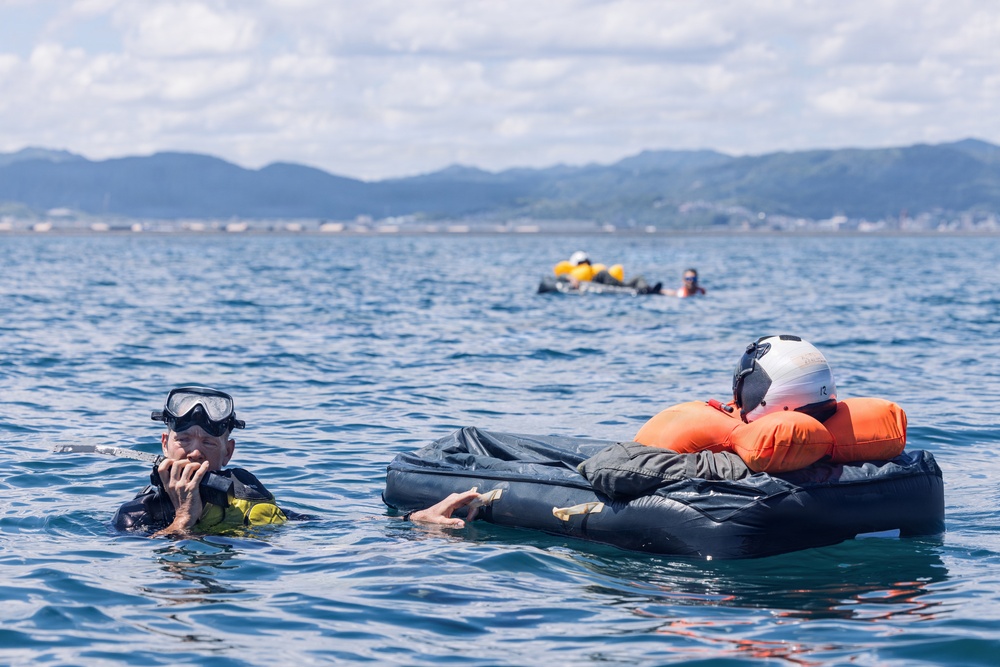 The image shows multiple people in open water engaged in what appears to be a water survival training or rescue exercise. In the foreground, one person wearing a diving mask and wetsuit is treading water, while another person in an orange life vest and white helmet is lying on a dark inflatable raft or survival platform. Additional participants and a yellow inflatable device are visible in the background.