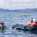 The image shows multiple people in open water engaged in what appears to be a water survival training or rescue exercise. In the foreground, one person wearing a diving mask and wetsuit is treading water, while another person in an orange life vest and white helmet is lying on a dark inflatable raft or survival platform. Additional participants and a yellow inflatable device are visible in the background.