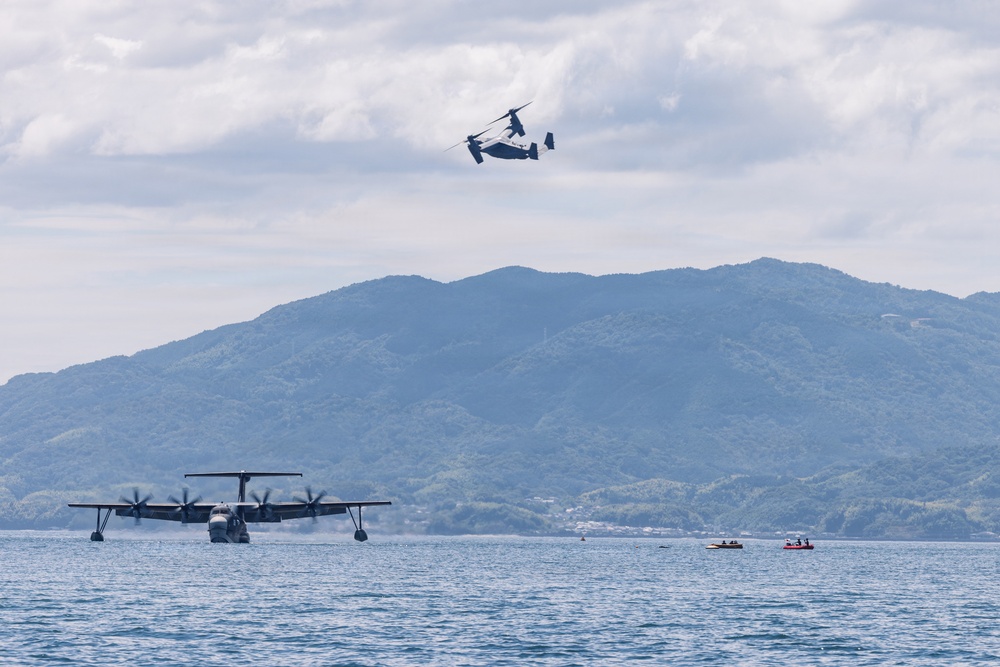 The image shows a maritime aviation scene over a calm bay with forested mountains in the background. A large four-engine flying boat or seaplane is taxiing or taking off on the water surface, while a tiltrotor aircraft (consistent with a V-22 Osprey type) flies overhead. Several small boats are visible on the water, suggesting this may be a military exercise or air show.