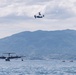 The image shows a maritime aviation scene over a calm bay with forested mountains in the background. A large four-engine flying boat or seaplane is taxiing or taking off on the water surface, while a tiltrotor aircraft (consistent with a V-22 Osprey type) flies overhead. Several small boats are visible on the water, suggesting this may be a military exercise or air show.