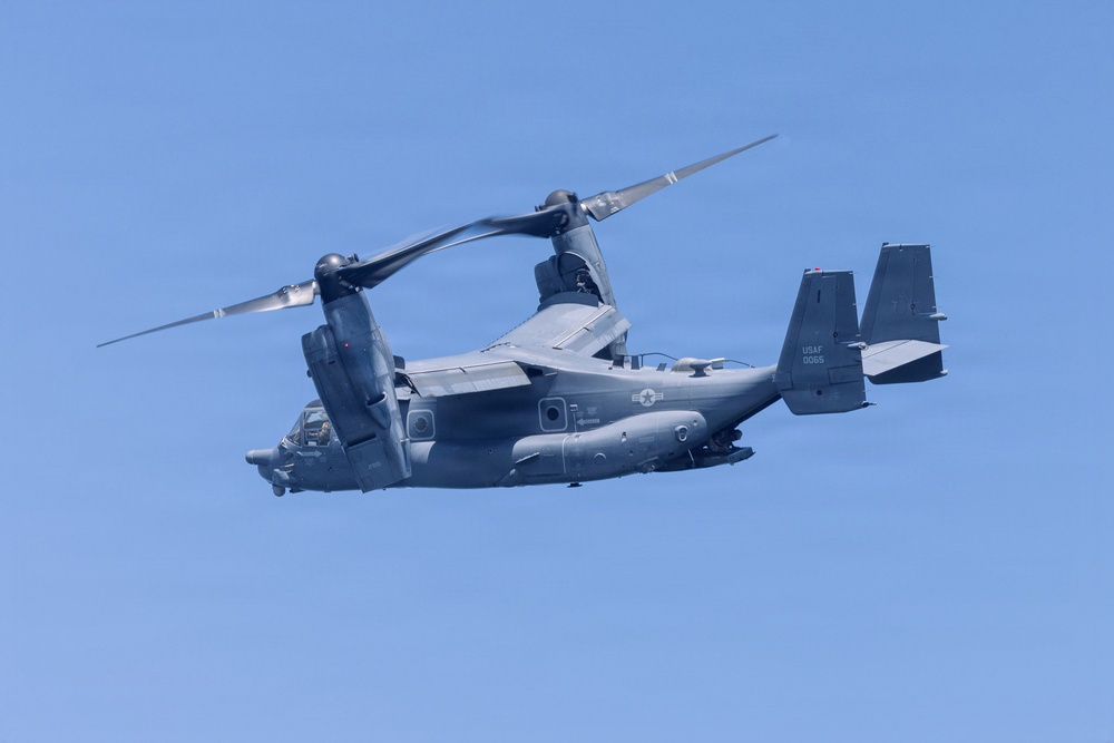 A United States Air Force CV-22 Osprey tiltrotor aircraft is captured in flight against a clear blue sky. The aircraft is painted in a dark gray/charcoal color scheme consistent with special operations use. Tail markings indicate it belongs to the USAF with the identifier '0065' visible on the tail fin.