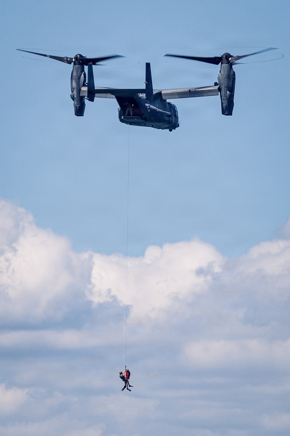 A dark-colored tiltrotor aircraft, consistent with a V-22 Osprey, is hovering in a partly cloudy blue sky. A hoist cable extends downward from the aircraft, with what appears to be two figures suspended at the end, suggesting a rescue or extraction operation. The image captures a mid-air personnel hoist or rescue demonstration.