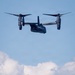 A dark-colored tiltrotor aircraft, consistent with a V-22 Osprey, is hovering in a partly cloudy blue sky. A hoist cable extends downward from the aircraft, with what appears to be two figures suspended at the end, suggesting a rescue or extraction operation. The image captures a mid-air personnel hoist or rescue demonstration.