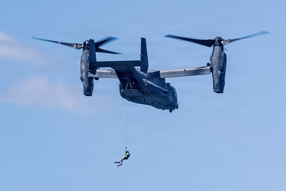 A military tiltrotor aircraft, identifiable as a Bell Boeing V-22 Osprey, is hovering in flight with its rotors in horizontal position. A person in what appears to be a rescue or combat diver configuration is suspended below the aircraft on a hoist or rappel line. This appears to be a search-and-rescue or special operations training exercise.