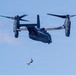 A military tiltrotor aircraft, identifiable as a Bell Boeing V-22 Osprey, is hovering in flight with its rotors in horizontal position. A person in what appears to be a rescue or combat diver configuration is suspended below the aircraft on a hoist or rappel line. This appears to be a search-and-rescue or special operations training exercise.