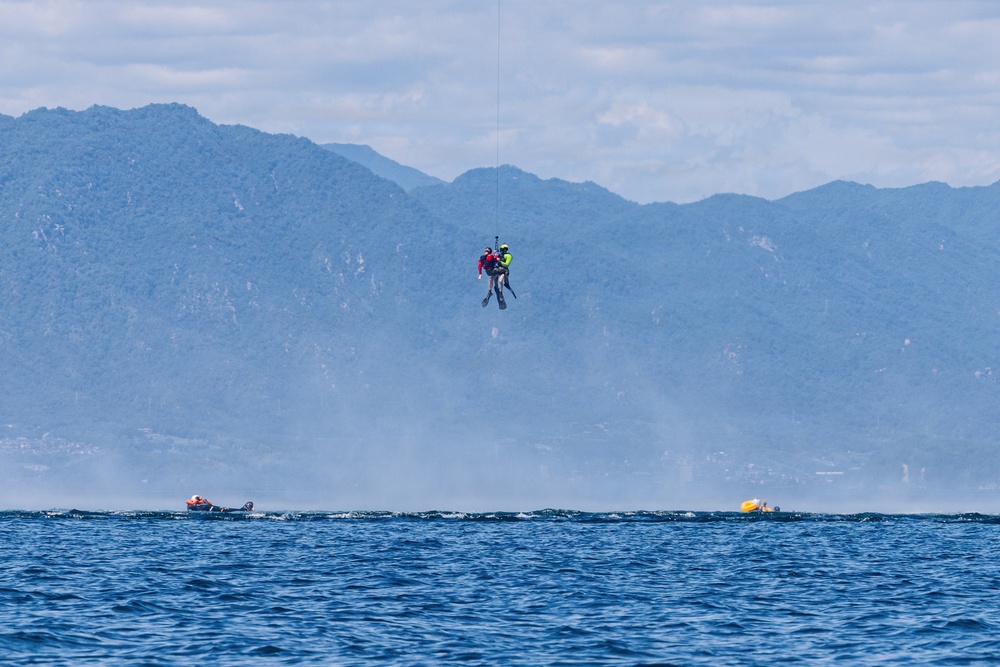 A helicopter hoist rescue operation is taking place over open water, with two individuals suspended mid-air on a cable — one appearing to be a rescuer in a yellow/green vest and the other a person being rescued in red. Two small watercraft or life rafts are visible on the water's surface below, with a mountainous, hazy landscape in the background. The scene suggests an active maritime search and rescue exercise or real emergency response.