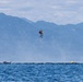 A helicopter hoist rescue operation is taking place over open water, with two individuals suspended mid-air on a cable — one appearing to be a rescuer in a yellow/green vest and the other a person being rescued in red. Two small watercraft or life rafts are visible on the water's surface below, with a mountainous, hazy landscape in the background. The scene suggests an active maritime search and rescue exercise or real emergency response.