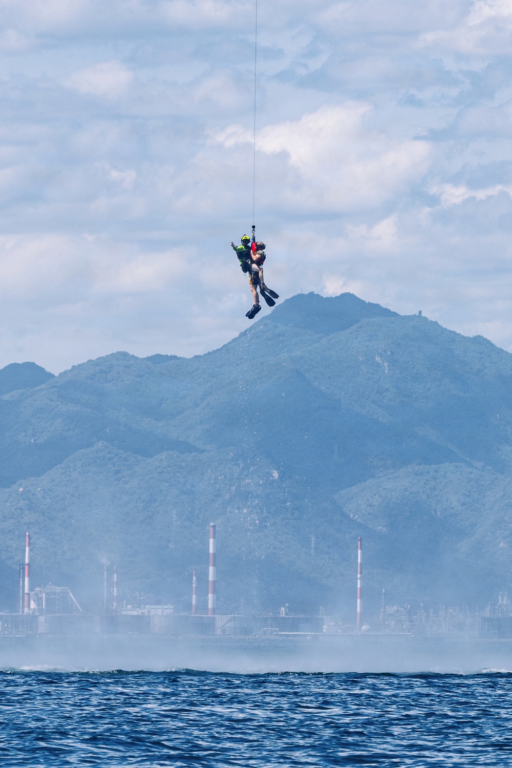 A helicopter rescue operation is underway over open water, with a rescue swimmer wearing a yellow helmet and diving fins hoisting what appears to be a survivor or casualty on a hoist cable. The scene is set against a backdrop of green mountainous terrain and an industrial facility with red-and-white striped smokestacks visible across the water. The image captures a mid-air extraction moment, suggesting a maritime search and rescue exercise or real operation.