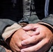 A close-up photograph of a pair of clasped, weathered hands resting on the lap of a person wearing tactical or military-style gear. The hands appear worn, dry, and calloused, suggesting physical labor or field work. The clothing includes a dark tactical vest with zippers and MOLLE-style webbing.