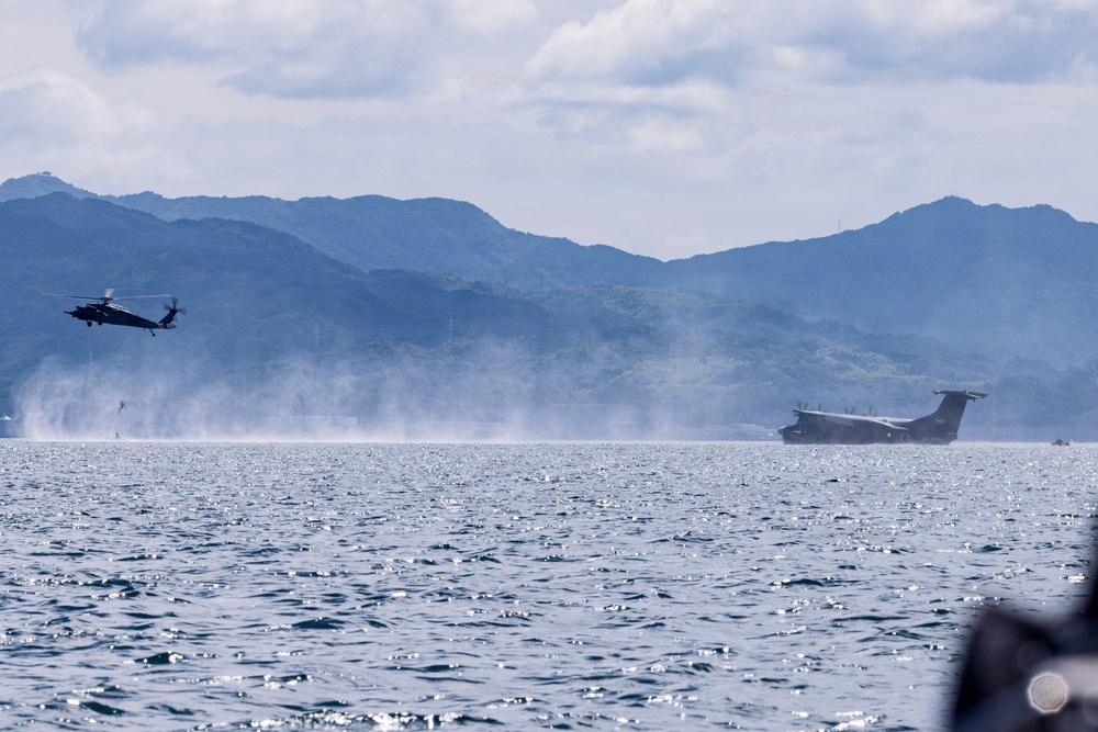 A military helicopter and a large flying boat or amphibious aircraft are operating over open coastal waters with mountainous terrain visible in the background. The flying boat appears to be on the water surface or taking off/landing, generating spray and mist. The scene suggests a joint military exercise or operational activity in a coastal maritime environment.