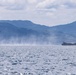 A military helicopter and a large flying boat or amphibious aircraft are operating over open coastal waters with mountainous terrain visible in the background. The flying boat appears to be on the water surface or taking off/landing, generating spray and mist. The scene suggests a joint military exercise or operational activity in a coastal maritime environment.