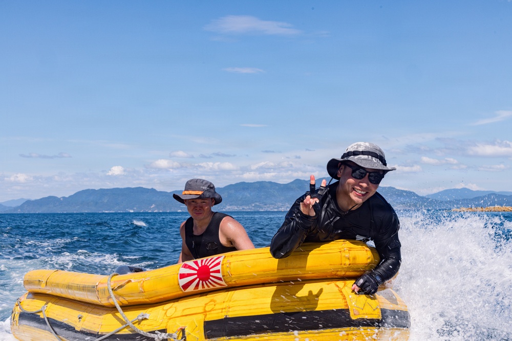 Two individuals are riding a yellow inflatable water raft or banana boat on open ocean waters. The person in the foreground is smiling and making a peace/victory hand gesture, wearing sunglasses and a bucket hat. Mountains and a coastline are visible in the background under a clear blue sky.