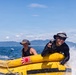 Two individuals are riding a yellow inflatable water raft or banana boat on open ocean waters. The person in the foreground is smiling and making a peace/victory hand gesture, wearing sunglasses and a bucket hat. Mountains and a coastline are visible in the background under a clear blue sky.