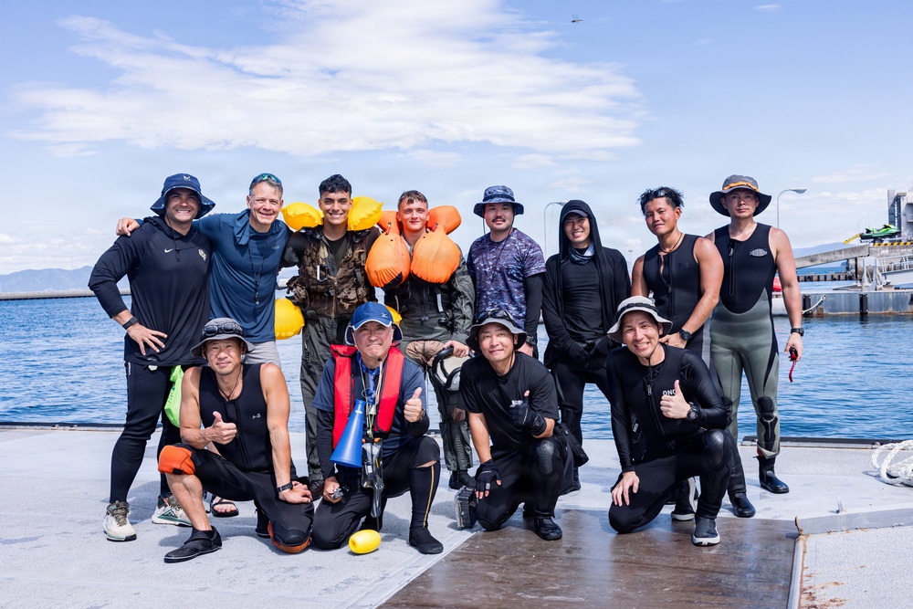 A group of approximately 13 individuals pose together on a waterfront dock or pier, appearing to be after a water-based activity. The group includes people wearing wetsuits, dive gear, life vests, and sun-protective clothing, suggesting a diving or water rescue training exercise. The setting appears to be a marina or harbor with calm blue water visible in the background.