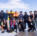 A group of approximately 13 individuals pose together on a waterfront dock or pier, appearing to be after a water-based activity. The group includes people wearing wetsuits, dive gear, life vests, and sun-protective clothing, suggesting a diving or water rescue training exercise. The setting appears to be a marina or harbor with calm blue water visible in the background.