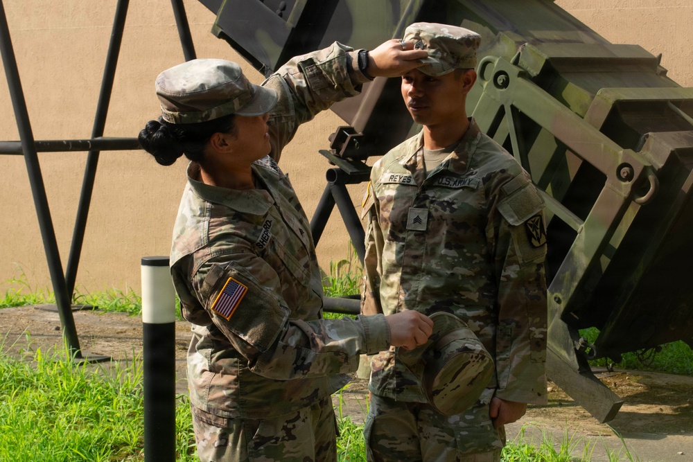 Two U.S. Army soldiers in OCP (Operational Camouflage Pattern) uniforms are engaged in what appears to be a promotion or pinning ceremony outdoors. One soldier (name tape reads 'SHEPARD') is placing or adjusting a patrol cap on the head of another soldier (name tape reads 'REYES'), who holds a cap in their hand. Military equipment, likely a Patriot missile system launcher or similar hardware, is visible in the background.