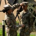 Two U.S. Army soldiers in OCP (Operational Camouflage Pattern) uniforms are engaged in what appears to be a promotion or pinning ceremony outdoors. One soldier (name tape reads 'SHEPARD') is placing or adjusting a patrol cap on the head of another soldier (name tape reads 'REYES'), who holds a cap in their hand. Military equipment, likely a Patriot missile system launcher or similar hardware, is visible in the background.