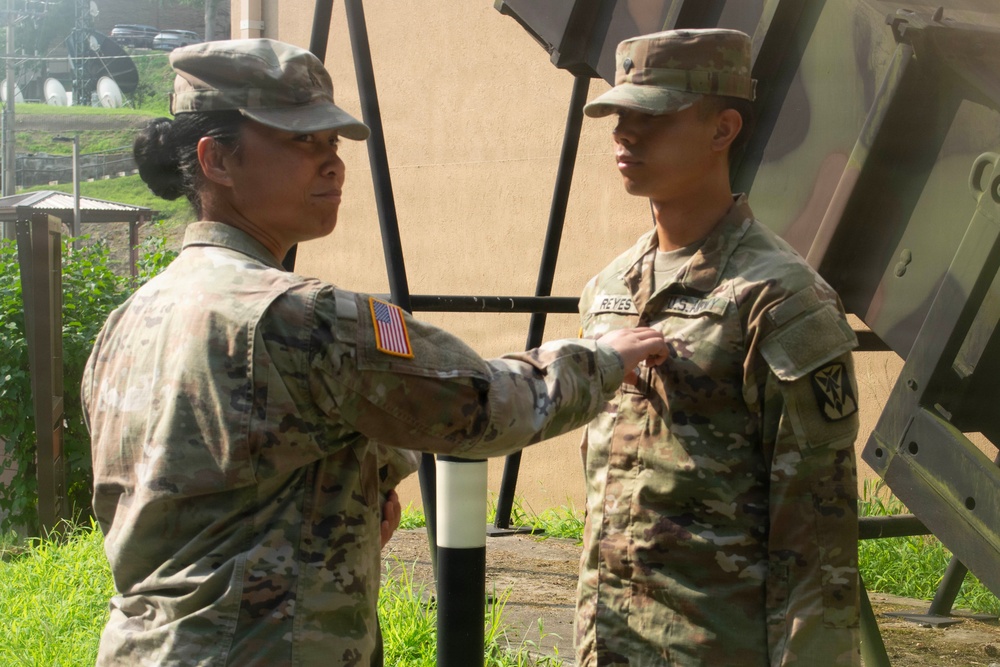 Two U.S. Army soldiers in OCP (Operational Camouflage Pattern) uniforms are engaged in what appears to be a pinning ceremony outdoors. The soldier on the left is reaching toward the chest area of the soldier on the right, whose name tape reads 'REYES.' Both wear U.S. Army patches and unit insignia consistent with an active-duty military setting.