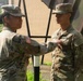 Two U.S. Army soldiers in OCP (Operational Camouflage Pattern) uniforms are engaged in what appears to be a pinning ceremony outdoors. The soldier on the left is reaching toward the chest area of the soldier on the right, whose name tape reads 'REYES.' Both wear U.S. Army patches and unit insignia consistent with an active-duty military setting.