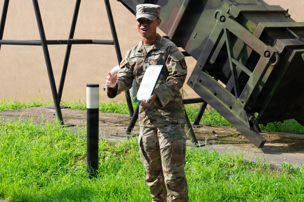 A U.S. Army soldier in OCP (Operational Camouflage Pattern) uniform, identified by the name tape 'REYES', stands outdoors holding a document or certificate. The soldier appears to be speaking or presenting, with one hand gesturing outward. The background includes a large military equipment structure, likely a Patriot missile launcher system, and a grassy area.