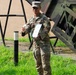 A U.S. Army soldier in OCP (Operational Camouflage Pattern) uniform, identified by the name tape 'REYES', stands outdoors holding a document or certificate. The soldier appears to be speaking or presenting, with one hand gesturing outward. The background includes a large military equipment structure, likely a Patriot missile launcher system, and a grassy area.