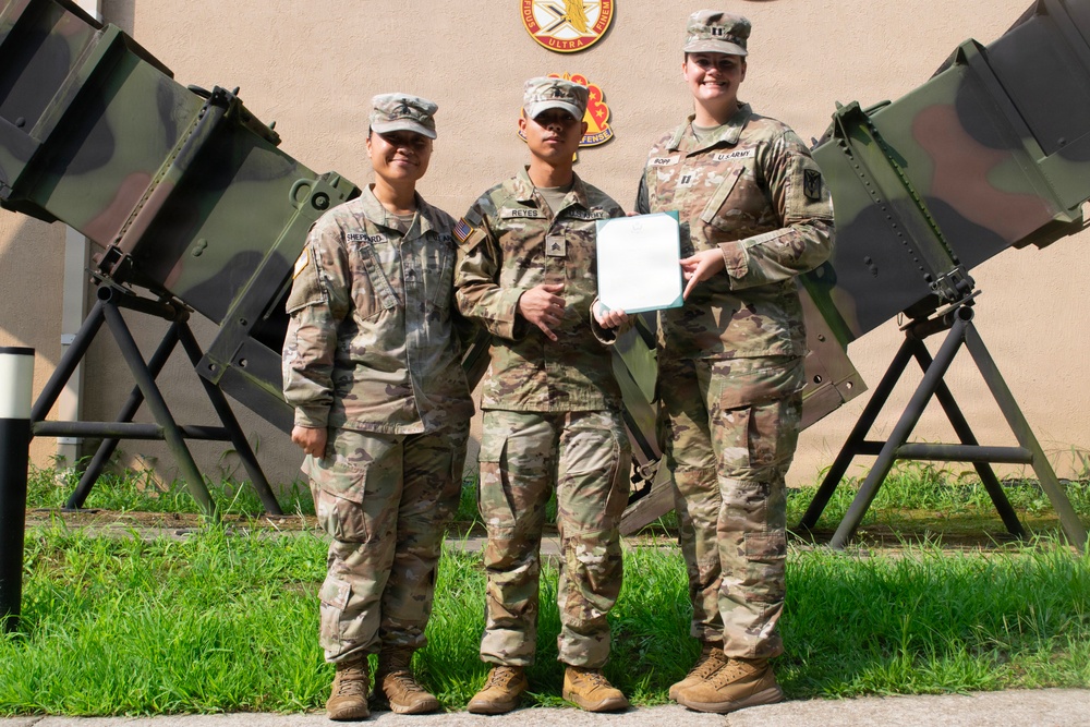 Three U.S. Army soldiers in OCP (Operational Camouflage Pattern) uniforms pose together outdoors in front of a military building. The center soldier is holding a document, suggesting a formal recognition or award ceremony. Military equipment, possibly Stinger missile launchers or similar air defense systems, is visible on both sides of the group.