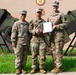 Three U.S. Army soldiers in OCP (Operational Camouflage Pattern) uniforms pose together outdoors in front of a military building. The center soldier is holding a document, suggesting a formal recognition or award ceremony. Military equipment, possibly Stinger missile launchers or similar air defense systems, is visible on both sides of the group.