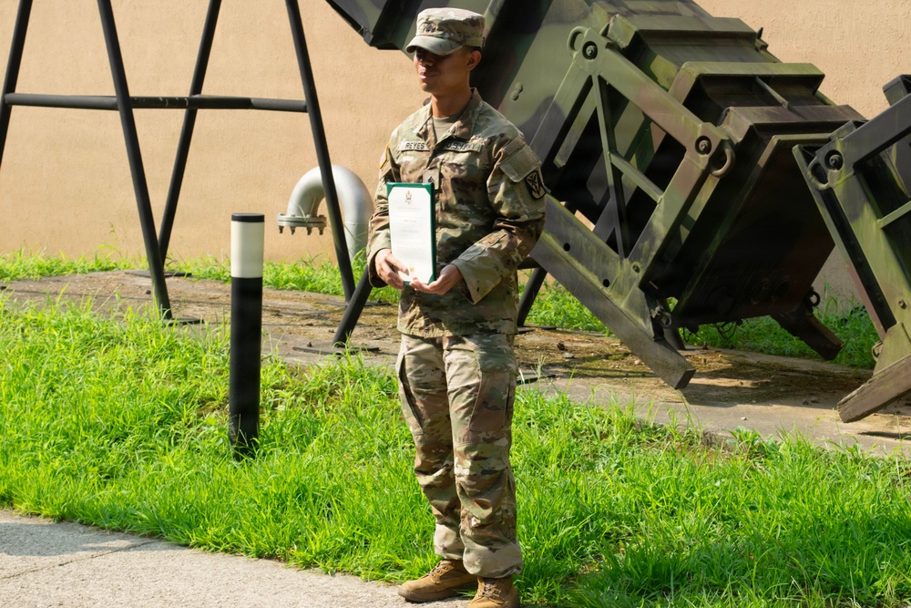 A U.S. Army soldier in Operational Camouflage Pattern (OCP) uniform stands outdoors holding what appears to be an award certificate in a green folder. The soldier's name tape reads 'REYES' and they are positioned in front of what appears to be military equipment, likely a Patriot missile launcher system. The setting suggests a military award or recognition ceremony conducted outdoors.
