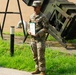 A U.S. Army soldier in Operational Camouflage Pattern (OCP) uniform stands outdoors holding what appears to be an award certificate in a green folder. The soldier's name tape reads 'REYES' and they are positioned in front of what appears to be military equipment, likely a Patriot missile launcher system. The setting suggests a military award or recognition ceremony conducted outdoors.