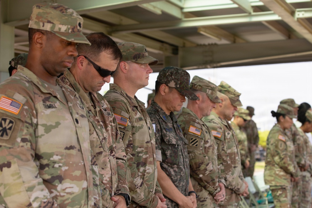 A group of military personnel in camouflage uniforms are standing in a line with heads bowed, suggesting a moment of prayer or solemn ceremony. The group includes both U.S. Army soldiers, identifiable by their OCP (Operational Camouflage Pattern) uniforms and American flag patches, and at least one soldier in what appears to be a Republic of Korea (ROK) Army uniform. The setting appears to be an outdoor covered structure, indicating a formal military event or ceremony.