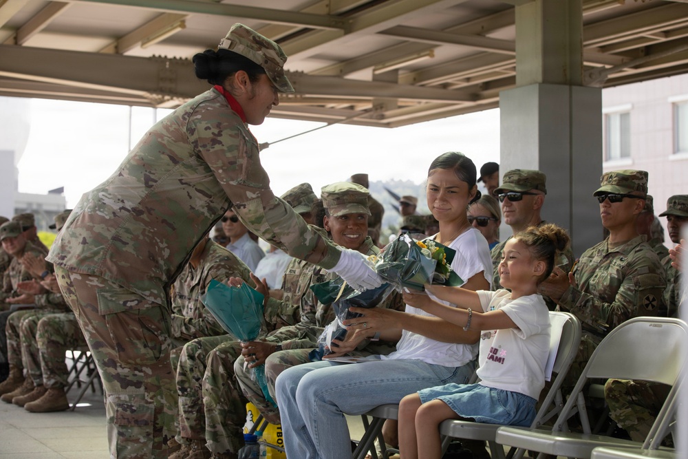 A military ceremony is taking place under a covered outdoor pavilion where a soldier in OCP (Operational Camouflage Pattern) uniform is presenting flowers to a young girl seated next to a woman in a white shirt. Multiple uniformed military personnel are seated in the background, some applauding. The scene suggests a formal recognition or welcome/farewell ceremony involving family members.