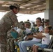 A military ceremony is taking place under a covered outdoor pavilion where a soldier in OCP (Operational Camouflage Pattern) uniform is presenting flowers to a young girl seated next to a woman in a white shirt. Multiple uniformed military personnel are seated in the background, some applauding. The scene suggests a formal recognition or welcome/farewell ceremony involving family members.