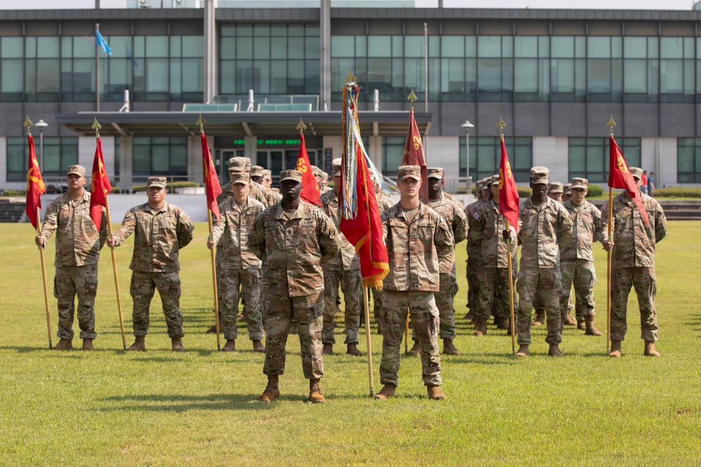 A formal military formation ceremony is taking place on a grass parade field in front of a large modern building. Soldiers in U.S. Army OCP (Operational Camouflage Pattern) uniforms are standing at attention in organized rows, with multiple guidon bearers holding red unit flags on wooden staffs. The central guidon bearer holds what appears to be a unit colors flag with gold fringe, suggesting this may be a change of command or activation ceremony.