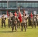 A formal military formation ceremony is taking place on a grass parade field in front of a large modern building. Soldiers in U.S. Army OCP (Operational Camouflage Pattern) uniforms are standing at attention in organized rows, with multiple guidon bearers holding red unit flags on wooden staffs. The central guidon bearer holds what appears to be a unit colors flag with gold fringe, suggesting this may be a change of command or activation ceremony.