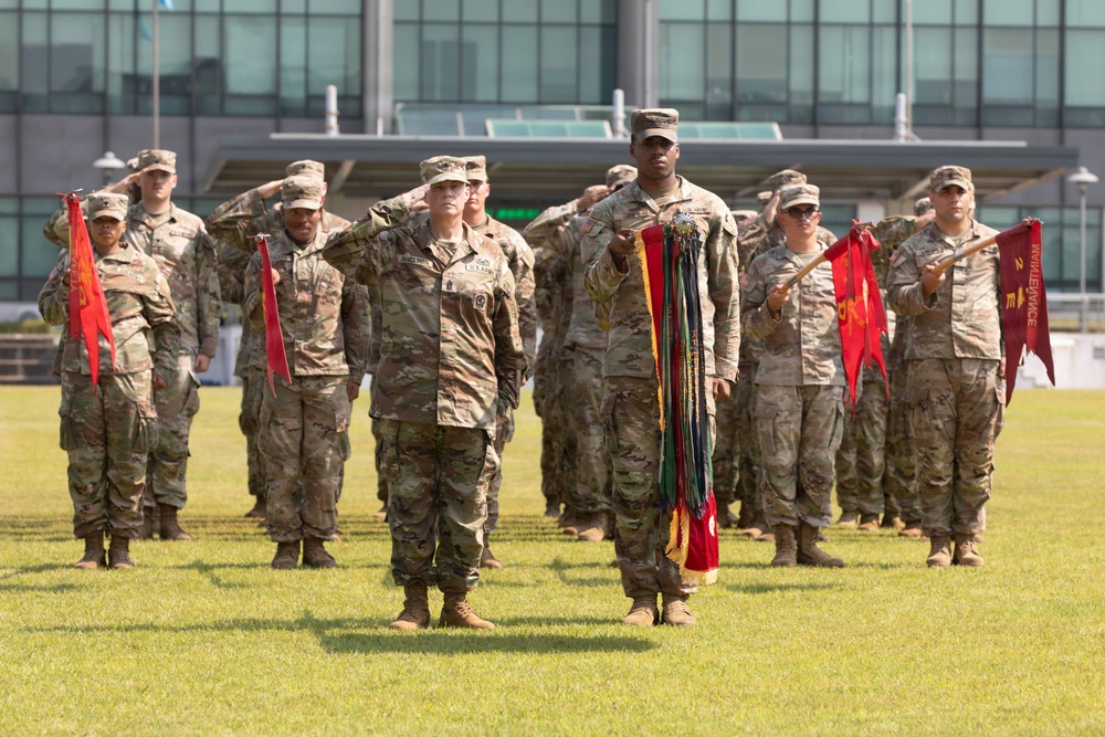 A formal U.S. Army military ceremony is taking place on a grass parade field in front of a modern glass building. Soldiers in Operational Camouflage Pattern (OCP) uniforms are standing in formation, with several rendering salutes and others holding red guidon flags with unit streamers. A central figure prominently holds a large collection of unit award streamers in multiple colors.