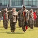 A formal U.S. Army military ceremony is taking place on a grass parade field in front of a modern glass building. Soldiers in Operational Camouflage Pattern (OCP) uniforms are standing in formation, with several rendering salutes and others holding red guidon flags with unit streamers. A central figure prominently holds a large collection of unit award streamers in multiple colors.