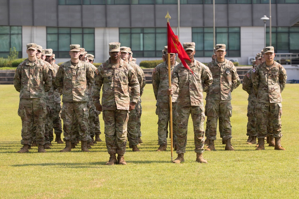 A formation of U.S. Army soldiers stands at parade rest on a grass field during what appears to be a military ceremony. The soldiers are wearing Operational Camouflage Pattern (OCP) uniforms and tan combat boots. A guidon bearer holds a red unit flag with a gold spearhead finial in the center of the formation.