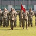 A formation of U.S. Army soldiers stands at parade rest on a grass field during what appears to be a military ceremony. The soldiers are wearing Operational Camouflage Pattern (OCP) uniforms and tan combat boots. A guidon bearer holds a red unit flag with a gold spearhead finial in the center of the formation.