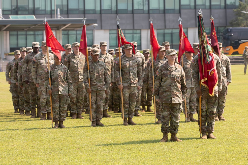A formal military formation ceremony featuring U.S. Army soldiers in Operational Camouflage Pattern (OCP) uniforms standing at attention on a grass field. Multiple guidon bearers are holding red unit flags/guidons with gold fringe, suggesting a change of command or unit activation ceremony. The event appears to take place at a military installation with a modern building visible in the background.