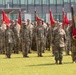 A formal military formation ceremony featuring U.S. Army soldiers in Operational Camouflage Pattern (OCP) uniforms standing at attention on a grass field. Multiple guidon bearers are holding red unit flags/guidons with gold fringe, suggesting a change of command or unit activation ceremony. The event appears to take place at a military installation with a modern building visible in the background.