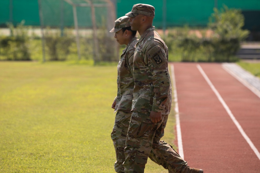 Two U.S. Army soldiers in Operational Camouflage Pattern (OCP) uniforms are walking side by side along the infield edge of a running track. Both are wearing patrol caps and appear to be in a formal or structured setting, possibly a military ceremony or inspection. The setting appears to be an outdoor athletic facility with a red rubberized track and green grass field.