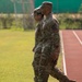Two U.S. Army soldiers in Operational Camouflage Pattern (OCP) uniforms are walking side by side along the infield edge of a running track. Both are wearing patrol caps and appear to be in a formal or structured setting, possibly a military ceremony or inspection. The setting appears to be an outdoor athletic facility with a red rubberized track and green grass field.