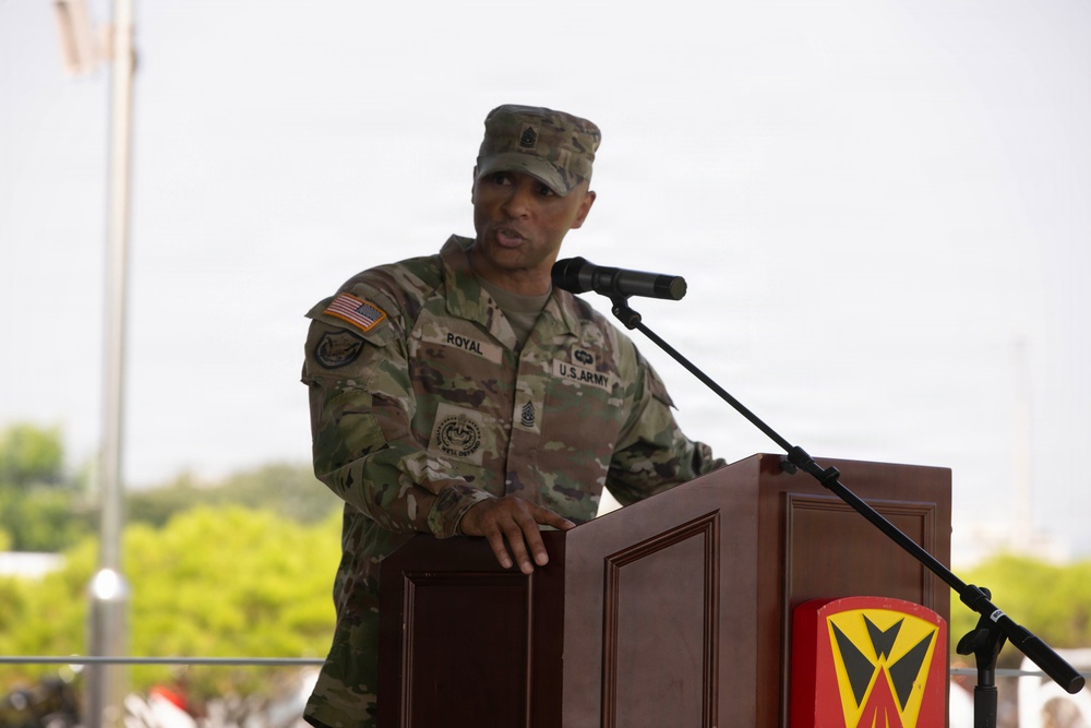 A U.S. Army soldier with the name tape 'ROYAL' is speaking at a wooden podium equipped with a microphone. The soldier is wearing Operational Camouflage Pattern (OCP) uniform with a U.S. flag patch, Sergeant Major rank insignia on the cap, and multiple unit patches. The podium displays a military unit insignia shield in red and yellow.