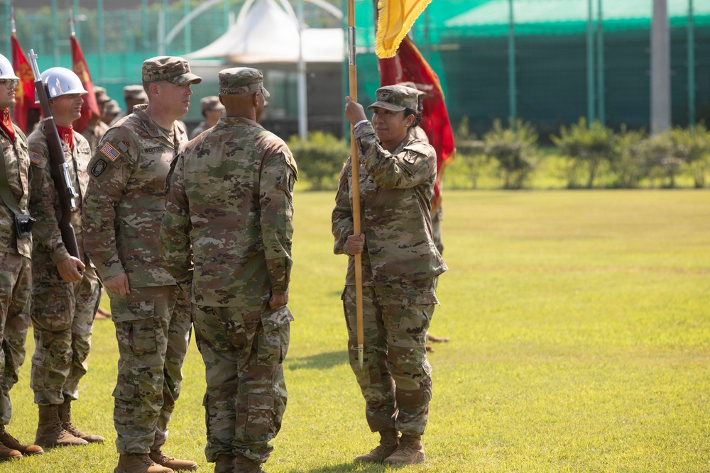 A U.S. Army military ceremony is taking place on a grass field, likely a change of command or activation ceremony. Soldiers in OCP (Operational Camouflage Pattern) uniforms are participating, with one soldier holding a yellow guidon flag while facing two other soldiers. Additional soldiers and color guard members with red flags are visible in the background.