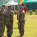 A U.S. Army military ceremony is taking place on a grass field, likely a change of command or activation ceremony. Soldiers in OCP (Operational Camouflage Pattern) uniforms are participating, with one soldier holding a yellow guidon flag while facing two other soldiers. Additional soldiers and color guard members with red flags are visible in the background.