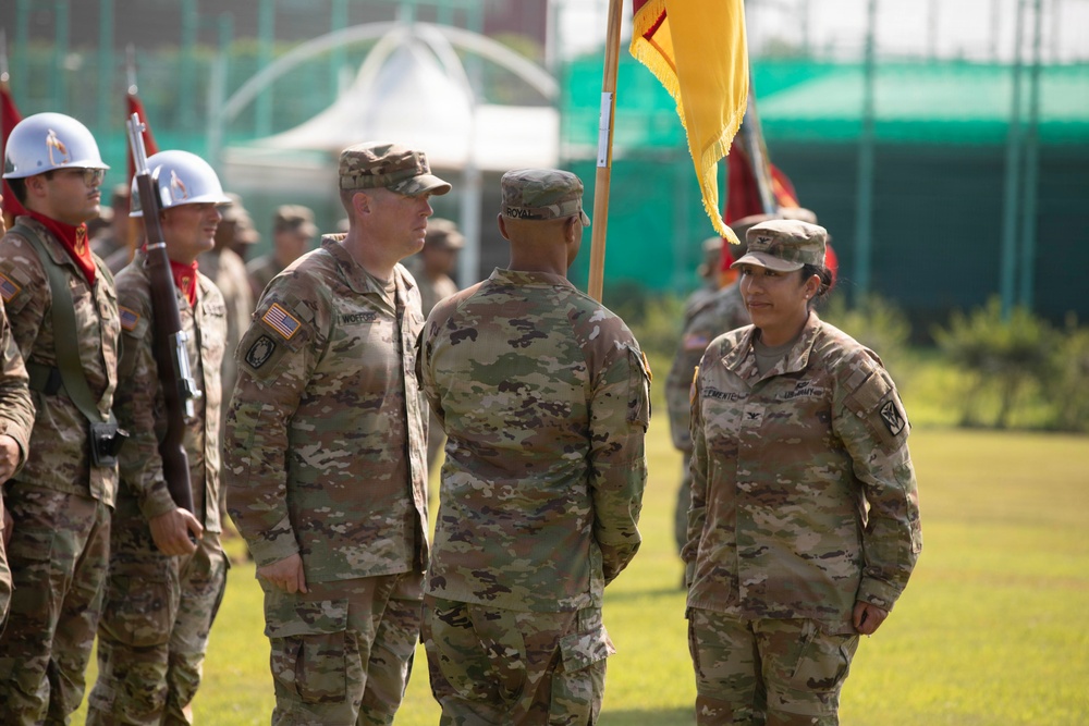 A military ceremony is taking place outdoors on a grass field, likely a change of command or unit activation ceremony. Several U.S. Army soldiers in OCP (Operational Camouflage Pattern) uniforms are gathered, with one soldier holding a yellow guidon flag. A ceremonial guard unit wearing white helmets and carrying rifles is visible in the background.